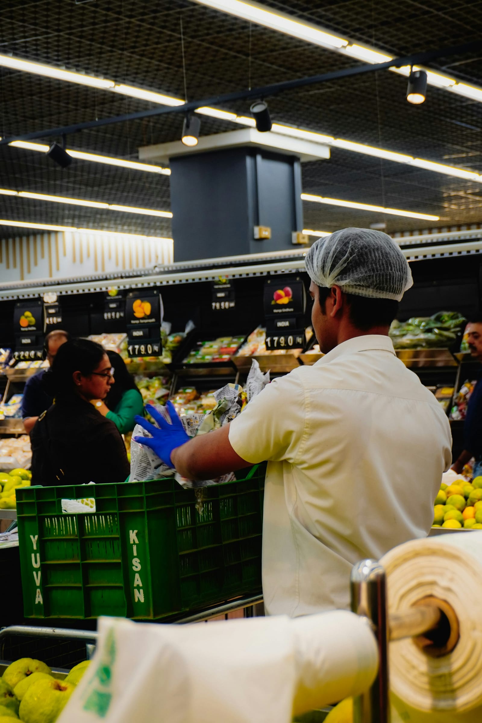 Funcionário de supermercado a repor fruta e legumes numa loja, com clientes ao fundo